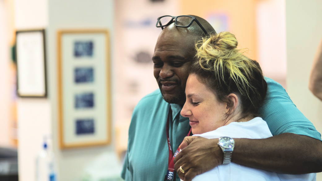Man hugging woman as another person holds a “Principal of the Year” award sign.