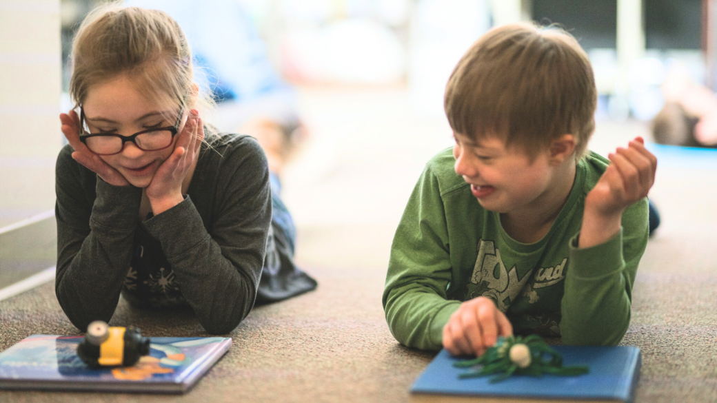 Two children lying on the floor smiling while playing with small toys on books.