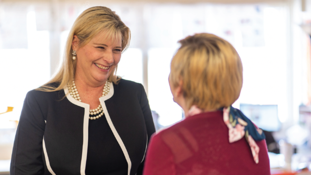 Smiling woman in a black suit talks with another woman in a red top.