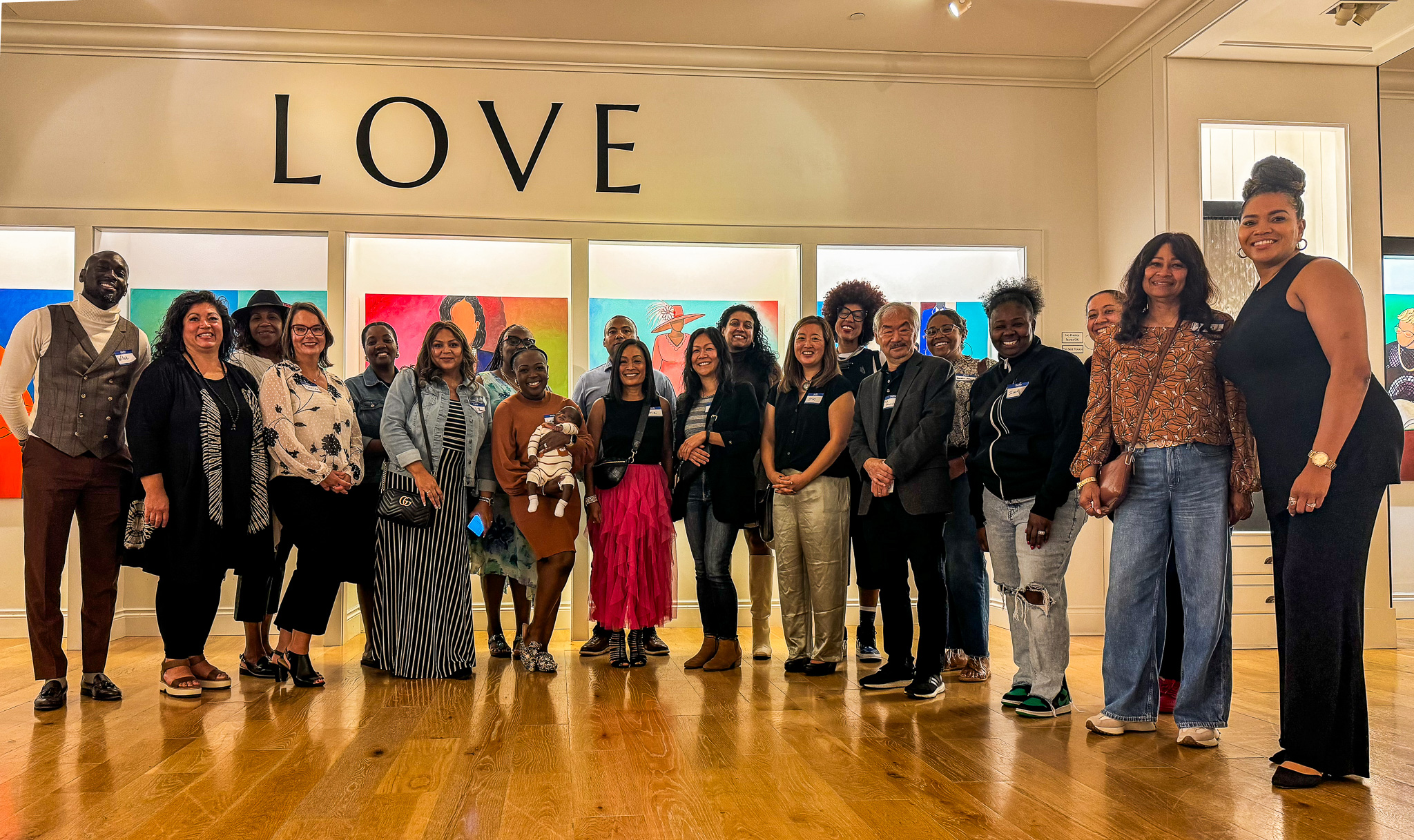 Group of people posing under a “LOVE” sign in front of colorful portrait artwork in a gallery setting.