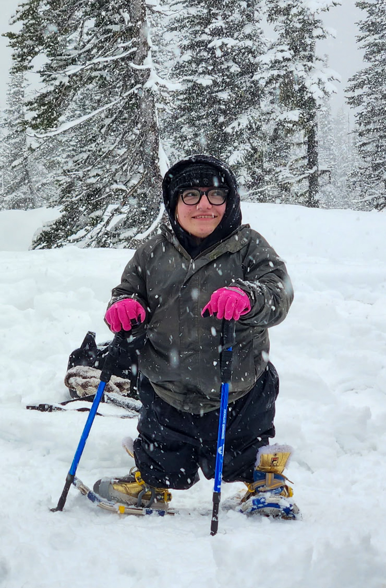 Person in winter gear snowshoeing in a snowy forest, using poles and wearing bright pink gloves.
