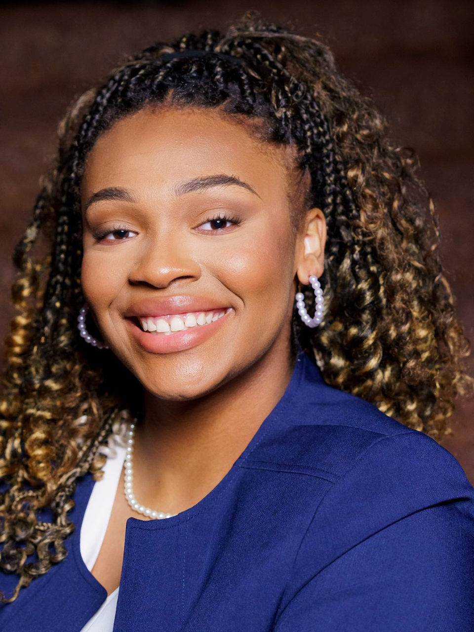 Young black woman with curly hair pulled back, wearing a blue blazer, white shirt, and pearl necklace.