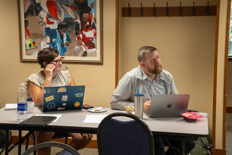 Man and women sitting at a table looking to the front of the room listening with laptops open.