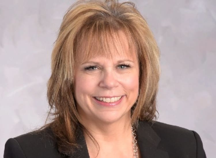Professional headshot of a smiling woman with shoulder-length light brown hair, wearing a black blazer and a statement necklace against a light gray background.