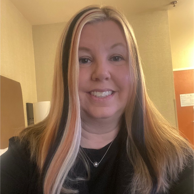 Smiling woman with long highlighted brown and blonde hair, wearing a black top and silver necklace, photographed indoors against a neutral background.