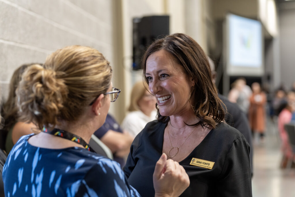 One white woman with a dress and nametag speaking to another white woman in a conference setting. 