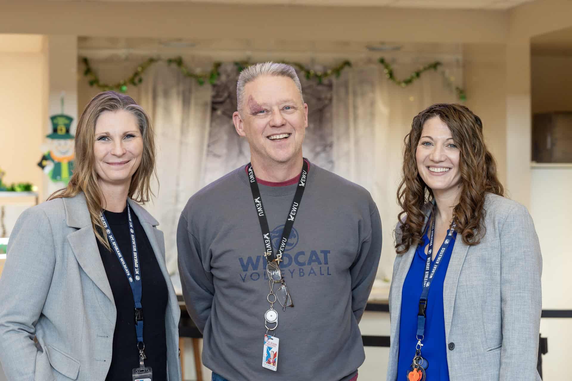 Three smiling adults wearing lanyards pose indoors; two women in blazers and one man in a sweatshirt.