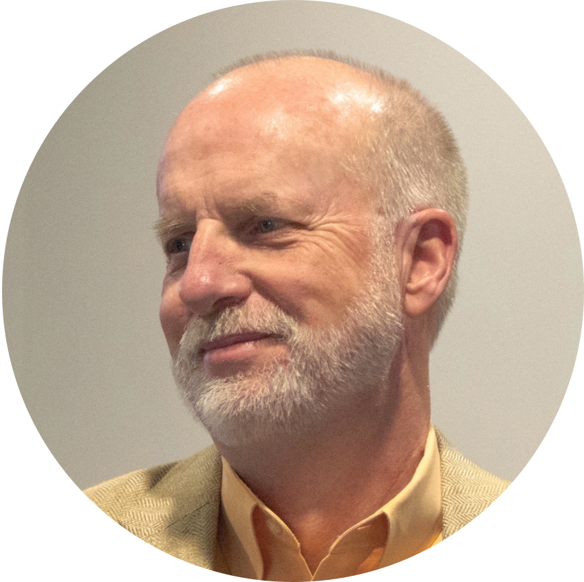 Close-up portrait of an older man with short gray hair and a neatly trimmed gray beard, wearing a tan blazer and yellow shirt, looking to the side.