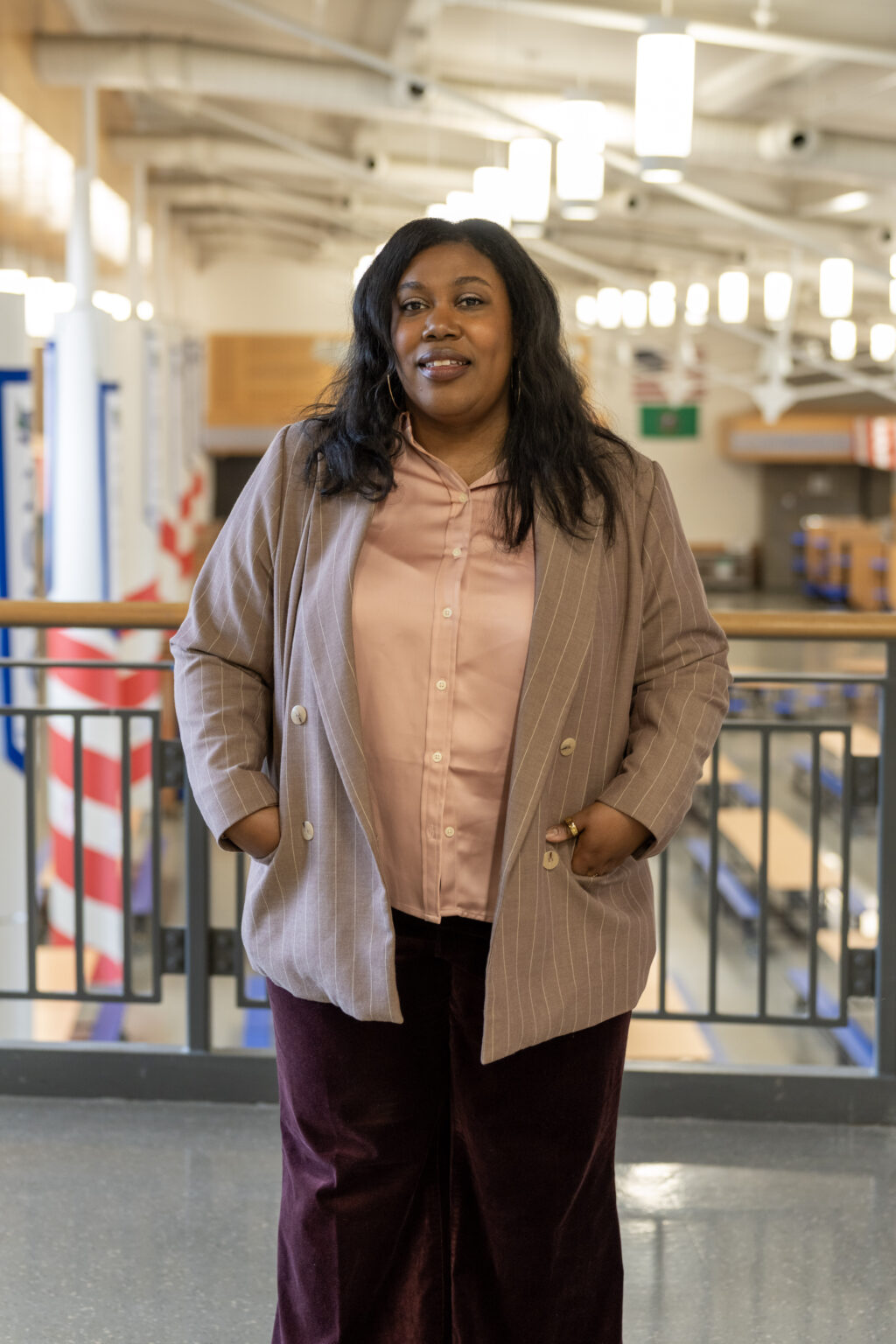 Portrait of a black woman standing indoors on an upper-level walkway, wearing a tan pinstriped blazer over a light pink blouse and dark pants, with hands in pockets and a calm, confident expression