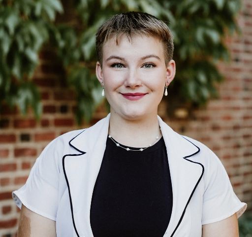 Selayna Rudolph poses with short hair wearing a white blazer over a black top, smiling at the camera, standing in front of a brick wall and leafy greenery