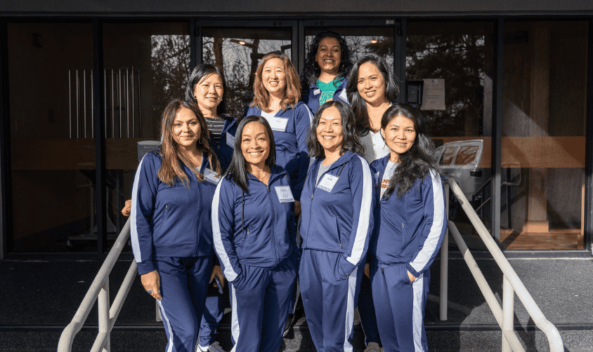Group of women in matching navy tracksuits smiling outside a building on a sunny day.