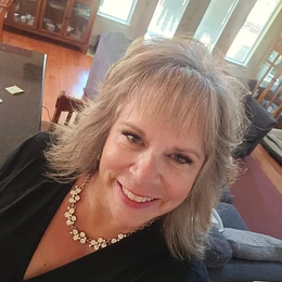 Smiling woman with shoulder-length gray hair, wearing a black top and floral necklace, seated indoors in an office or meeting space.