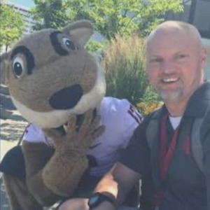 A smiling bald man with a gray beard poses outdoors beside a large brown animal mascot wearing a purple shirt.avatar small