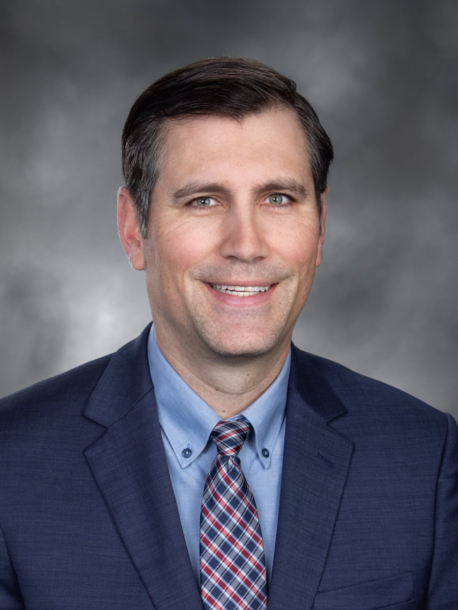 Portrait of Representative Steve Bergquist, Washington State legislator for the 11th District, wearing a blue suit, light blue shirt, and plaid tie against a gray studio background.