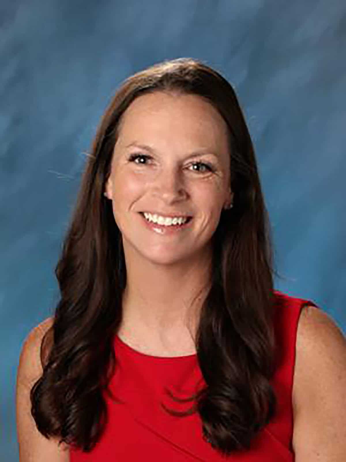 Smiling woman with long, wavy brown hair wearing a red sleeveless dress against a blue gradient background.