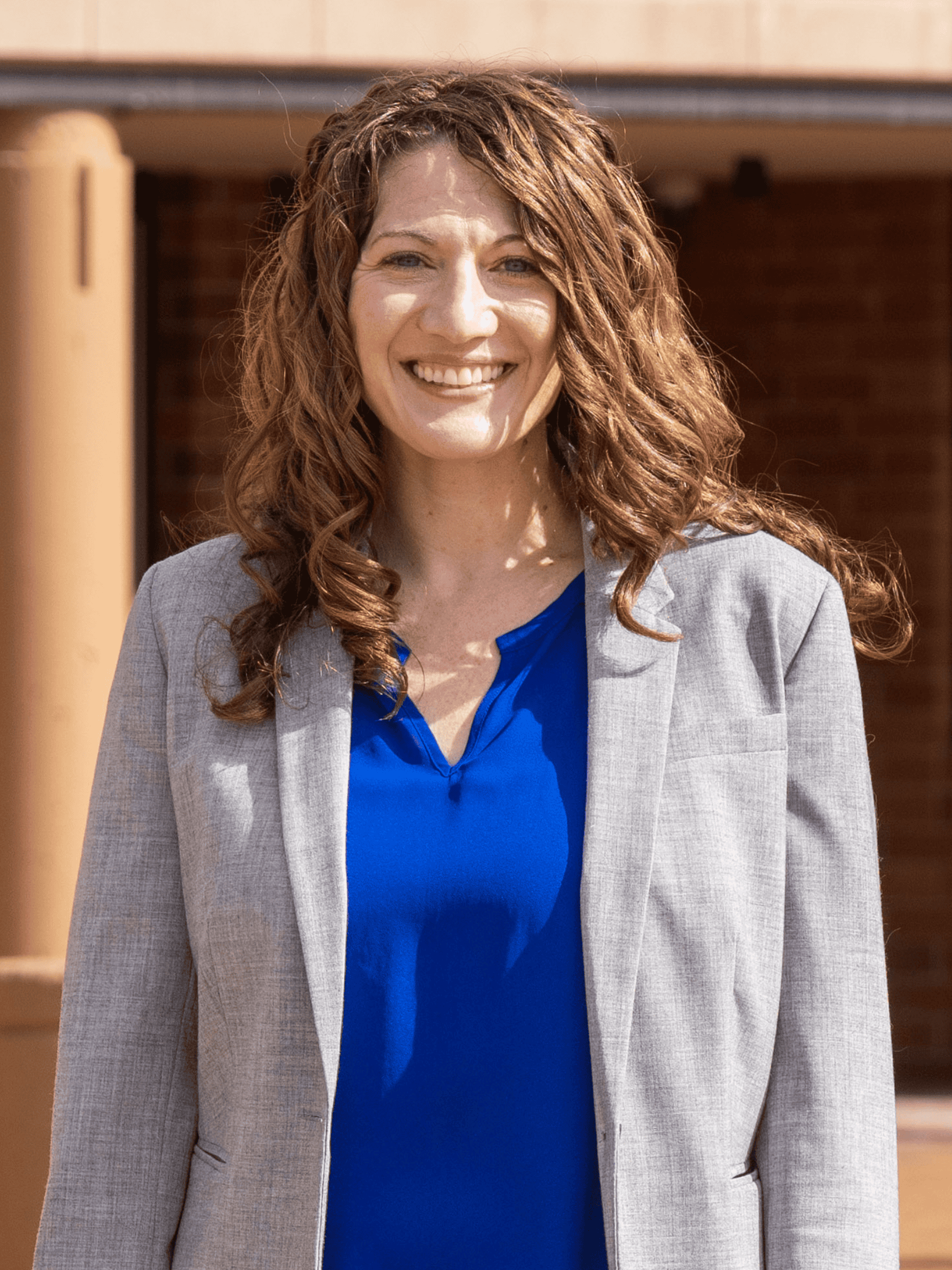 A smiling woman with curly hair wears a gray blazer and royal blue blouse, standing outdoors in front of a building with brick and tan columns.