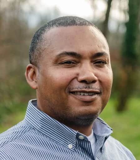 Smiling man in a blue and white striped shirt standing outdoors with a blurred green background, showcasing a friendly demeanor.