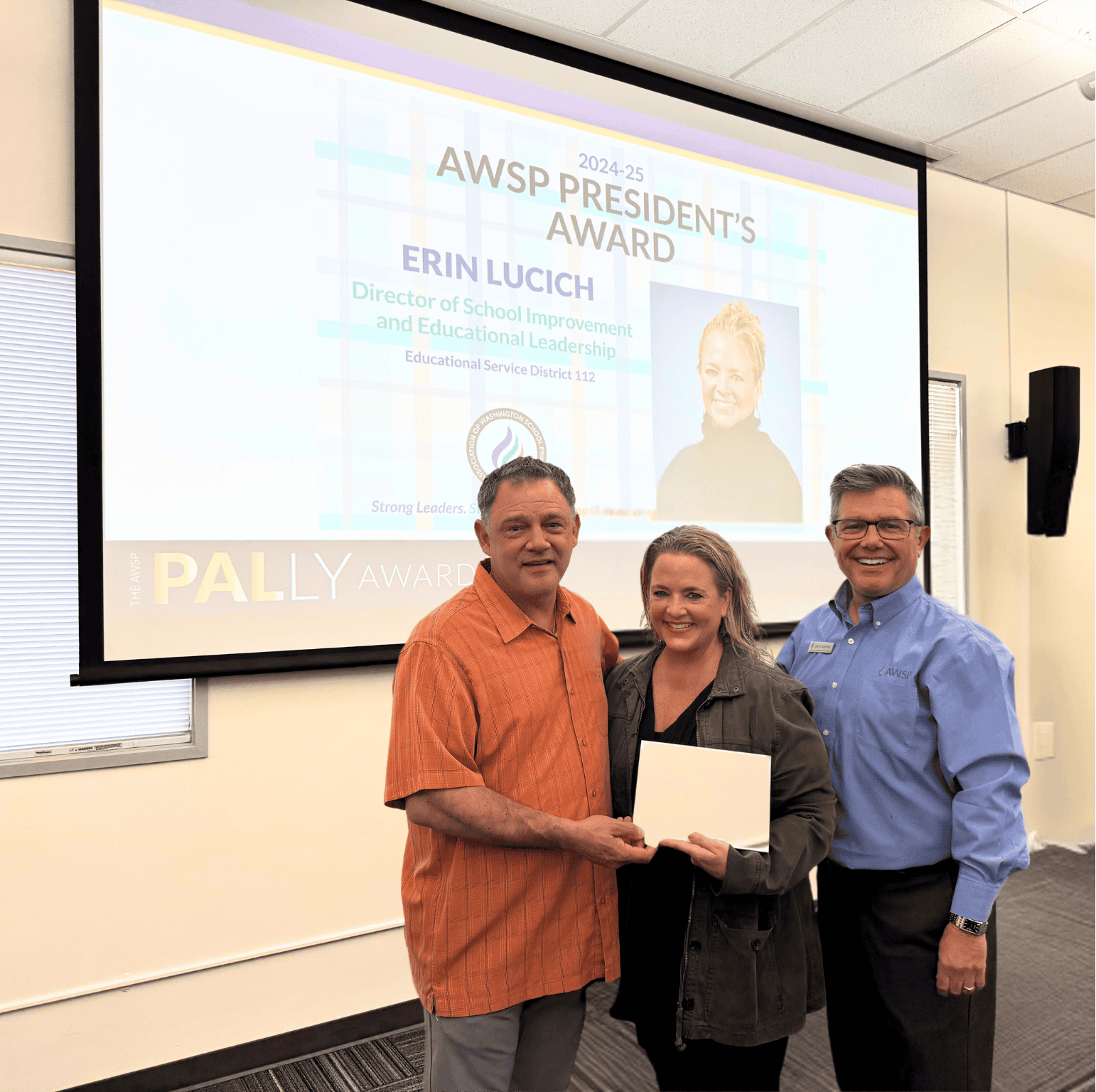 Three people pose indoors with an award; presentation slide for Erin Lucich is displayed in the background.