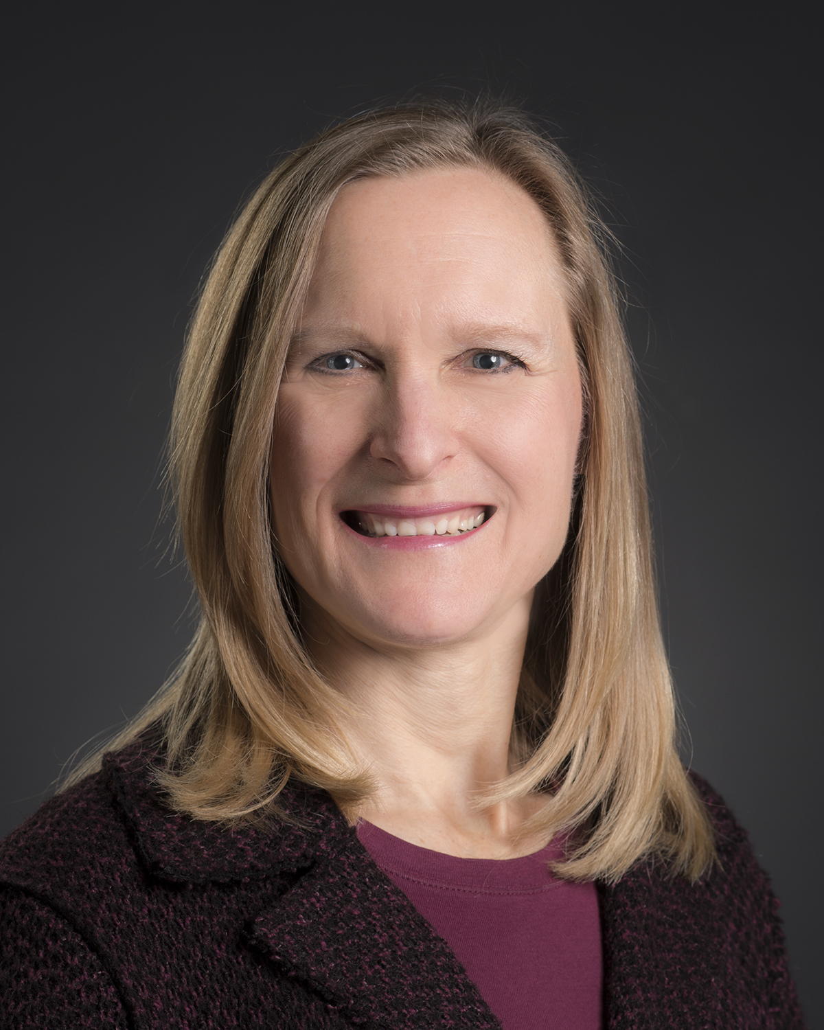 Professional headshot of a smiling woman with shoulder-length blonde hair, wearing a dark burgundy textured blazer over a plum-colored top. She is facing the camera against a dark gray backdrop with soft studio lighting. The portrait is framed from the chest up and conveys a friendly, approachable demeanor.