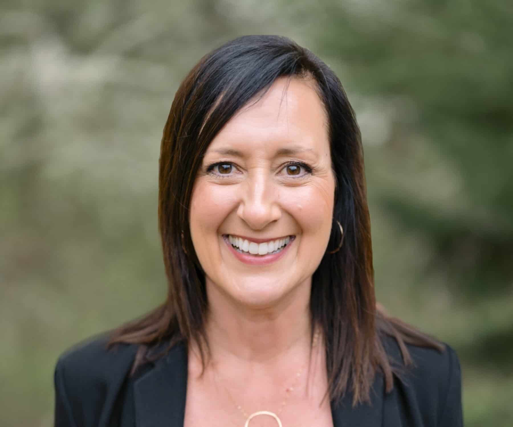 A smiling woman with shoulder-length dark hair, wearing a black blazer and gold necklace, standing outdoors with a blurred green background
