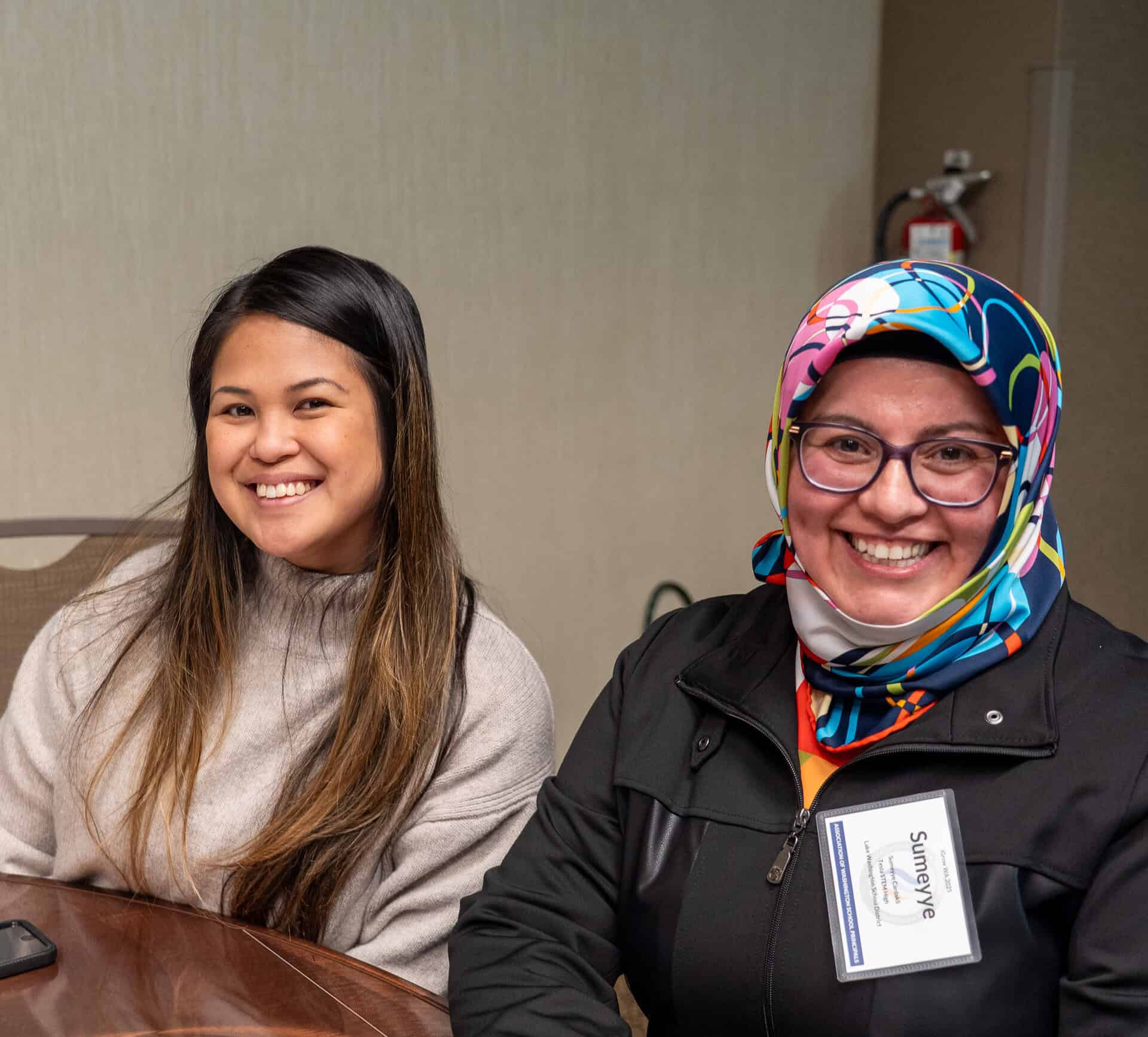 Two women smiling at a table, one in a sweater, the other in a colorful hijab and name tag.