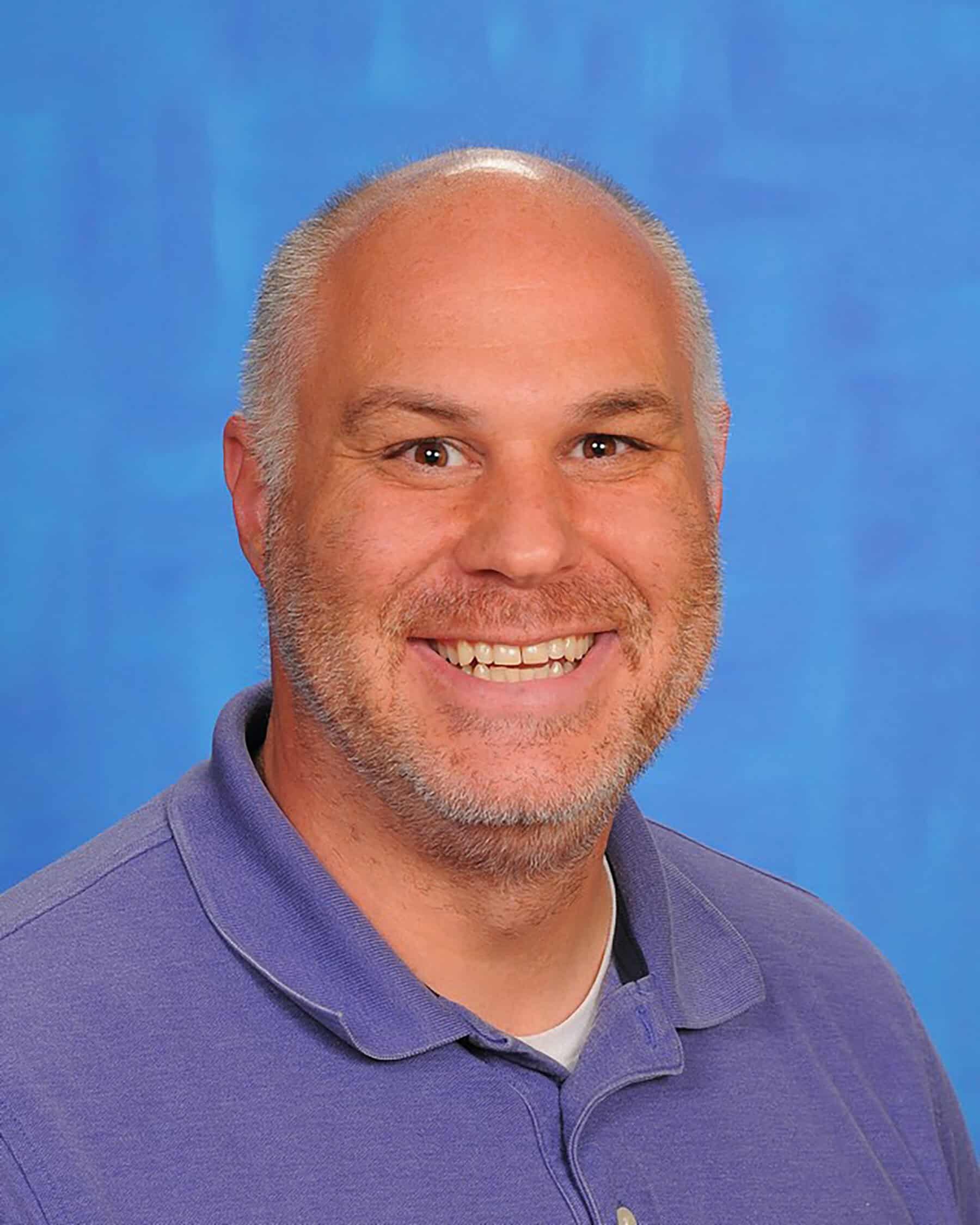 Smiling bald man with light stubble wearing a purple polo shirt against a blue backdrop.