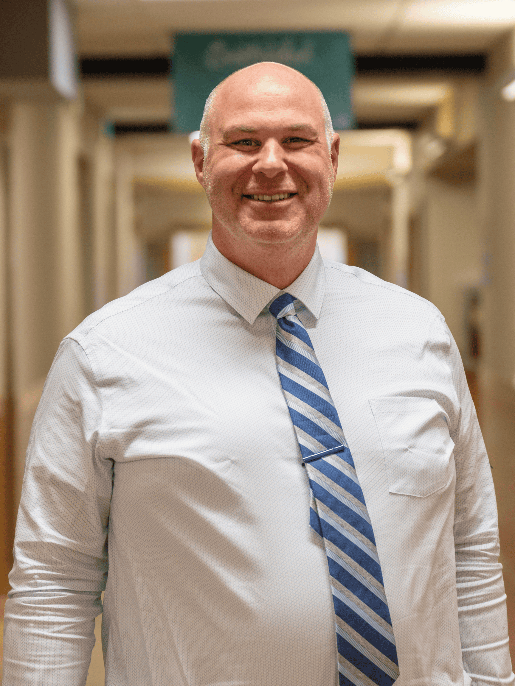 A smiling bald man in a white dress shirt and blue striped tie stands in a hallway with soft lighting and a blurred background.