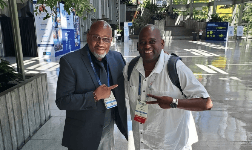 Two smiling Black men at a conference, pointing at each other in a bright, modern atrium with banners behind.
