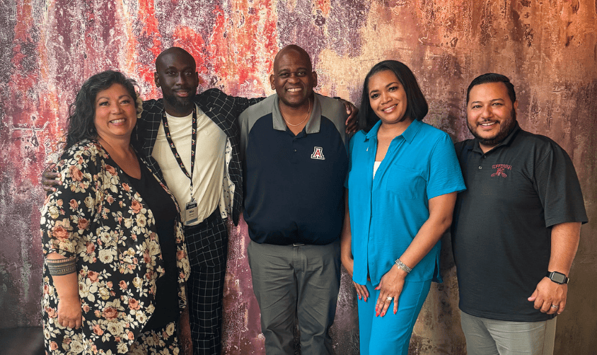 Group of five smiling adults posing together in front of a textured, multicolored wall.