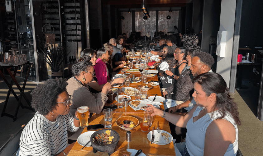 Large group dining together at a long restaurant table, enjoying food and conversation in warm sunlight.