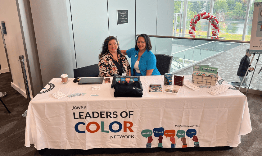 Two women sit at a “Leaders of Color” info table with brochures and swag at a bright indoor event.