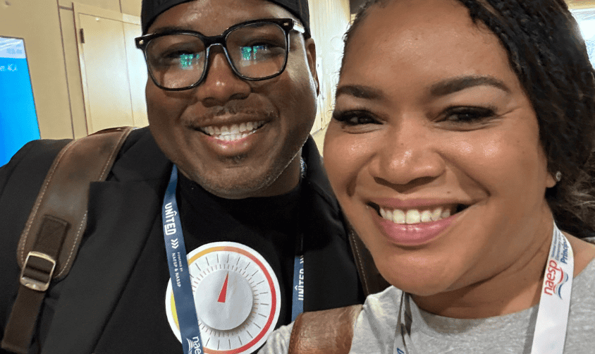 Two smiling Black people wearing lanyards take a close-up selfie at a conference.