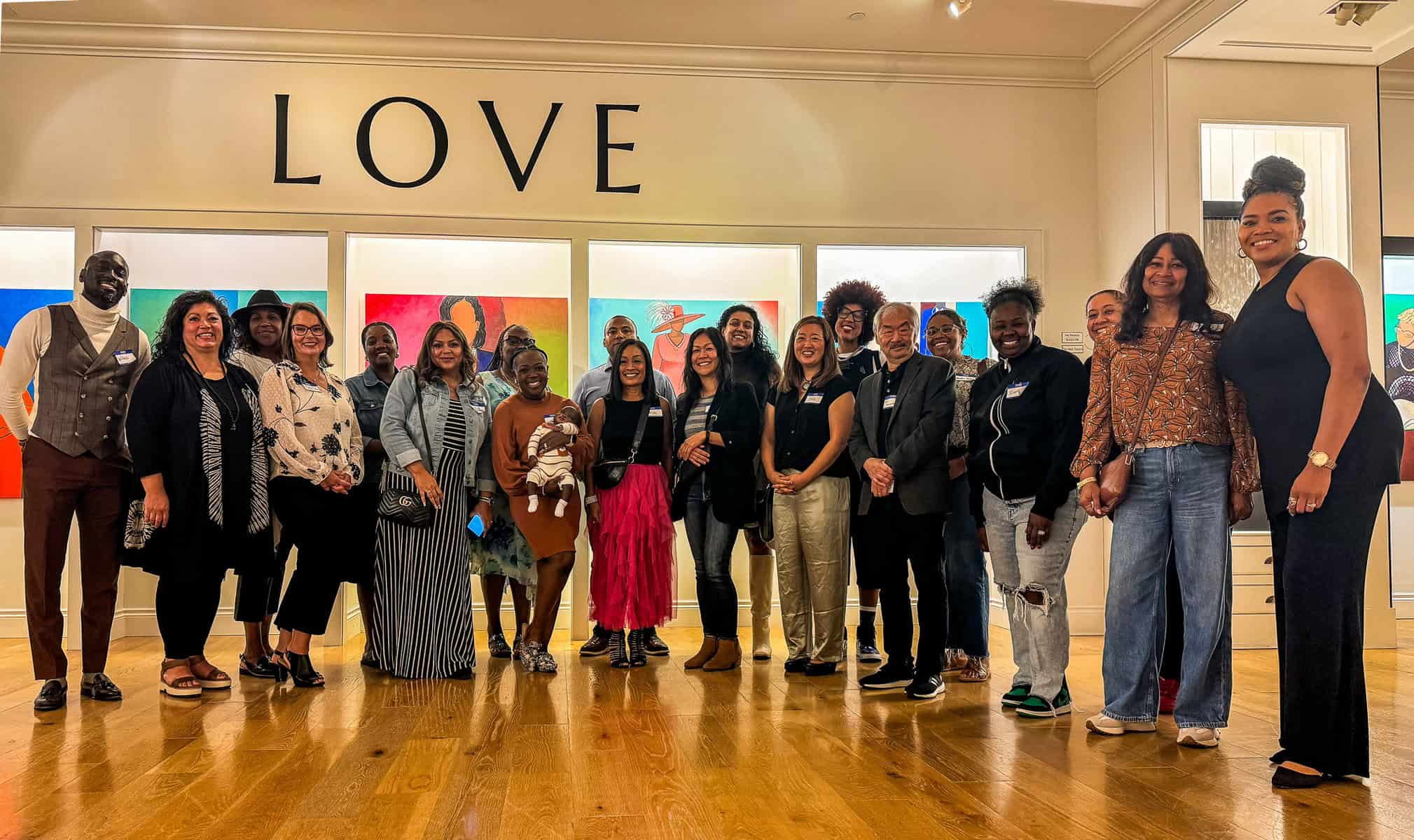 Group of people posing under a “LOVE” sign in front of colorful portrait artwork in a gallery setting.