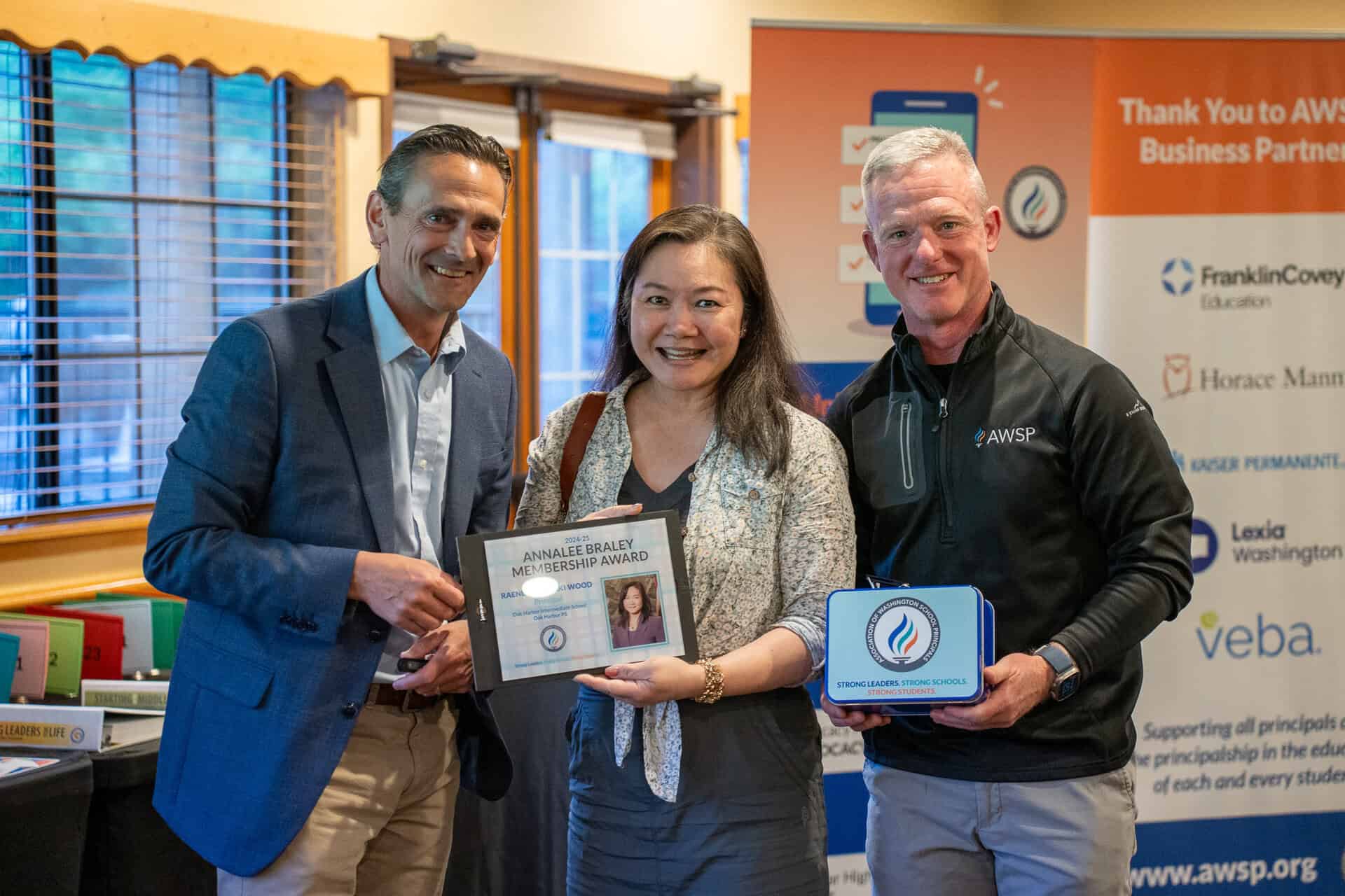 Three people smiling indoors, holding an award plaque and a blue AWSP-branded lunchbox.