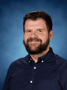 Smiling man with short brown hair and a beard, wearing a navy blue button-up shirt, against a blue backdrop.