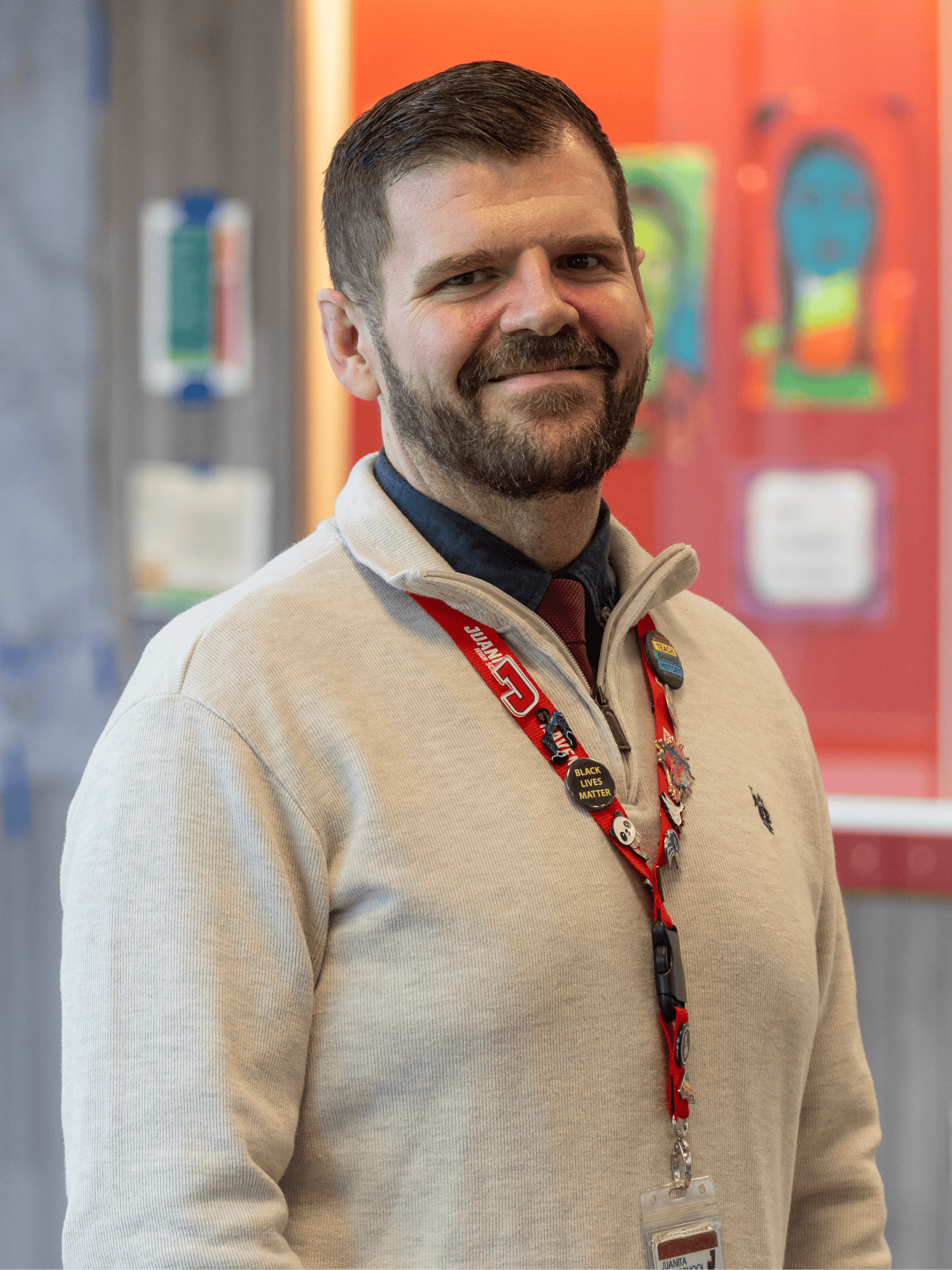 A smiling man with a beard wears a cream sweater, red lanyard with pins, and name badge, standing indoors with colorful student artwork in the blurred background.
