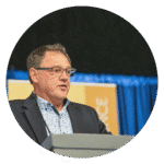 Speaker presenting at a conference, wearing glasses and a suit, with a blue backdrop and a podium.